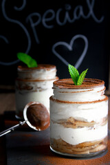 Two jars filled with tiramisu with cocoa powder on top and mint leaves as decoration on a chalkboard background. Dark food concept.