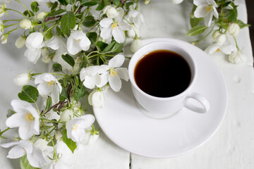 White cup of morning espresso with a blossoming apple tree branch on a wooden surface of a table with reflections.