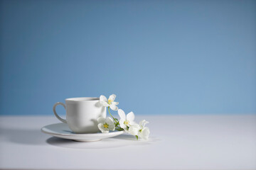 White cup of morning espresso with a blossoming apple tree branch on a stone surface of a table with reflections opposite blue background.