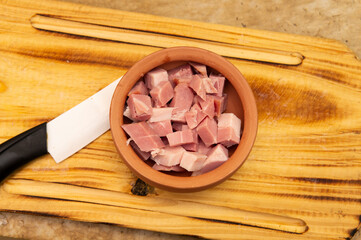 Close up view of a small bowl with bacon chopped in dices inside next to a white ceramic knife. Placed on top of a wooden cutting board. Taken indoor under a soft white light.