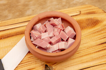 Close up view of a small bowl with bacon chopped in dices inside next to a white ceramic knife. Placed on top of a wooden cutting board. Taken indoor under a soft white light.