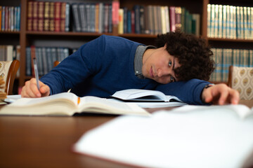 A tired curly-haired student in a grey-collared blue sweater, lay on his arm tired of homework and looking at the camera in the library