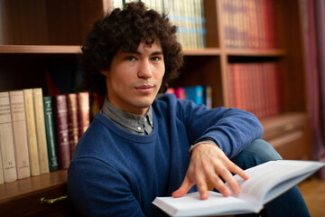 Portrait of a handsome curly boy student looking at the camera in a gray shirt, and blue sweater with an open book against the background of bookshelves in the library. High quality photo