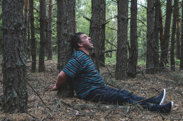 Naklejka na ściany i meble A hostage in the woods. A man in a blue t-shirt and trousers is sitting on the ground tied to a tree in the forest calling for help. Victim of an attack. Horizontal photo.