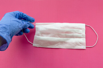 A man's hand in a blue medical glove holds a medical mask on a pink background. Selective focus.