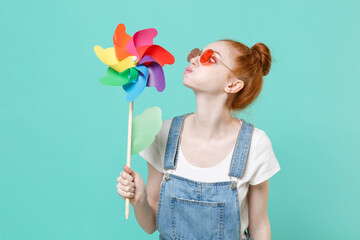 Funny young readhead girl in casual denim clothes eyeglasses posing isolated on blue turquoise wall background studio portrait. People lifestyle concept. Mock up copy space. Blowing on toy windmill.