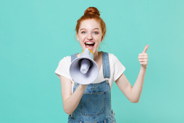 Funny young readhead girl in casual denim clothes posing isolated on blue turquoise wall background studio portrait. People lifestyle concept. Mock up copy space. Scream in megaphone showing thumb up.
