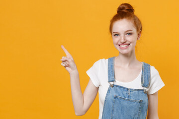 Smiling young readhead girl in casual denim clothes white t-shirt isolated on yellow background studio portrait. People emotions lifestyle concept. Mock up copy space. Pointing index finger aside up.