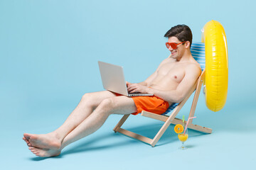 Smiling young man guy in orange shorts glasses sit on deck chair isolated on blue background studio. People summer vacation rest lifestyle concept. Mock up copy space. Working on laptop pc computer.