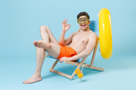 Funny young man guy in orange shorts swimming goggles sit on deck chair isolated on blue wall background studio. People summer vacation rest lifestyle concept. Mock up copy space. Showing OK gesture.