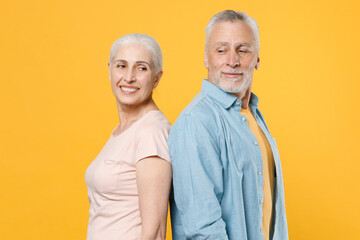 Side view of smiling elderly gray-haired couple woman man in casual clothes isolated on yellow background. People lifestyle concept. Mock up copy space. Standing back to back looking at each other.