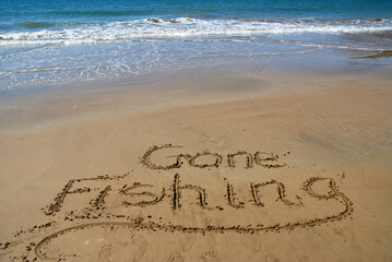 Gone fishing written in sand on the beach with surf and blue sea beyond.