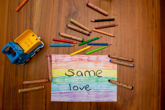 Child painting on a wooden table reading; "same love" on a rainbow flag, colorful crayons and a toy truck around it