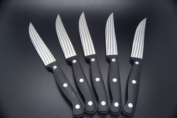 Set of five silver steak knives with black handles on black background