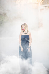 Beautiful woman model with long blond hair in a black evening dress with open neckline decoration on the neck, the bracelet on arm and pink lips standing in the smoke from the bombs on the background