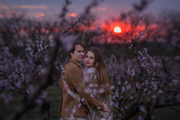 Obraz premium Young couple in love hug and kiss on the background of blooming gardens during sunset. A beautiful girl looks at the camera, a man kisses her on the lips and forehead. Wedding anniversary celebration,