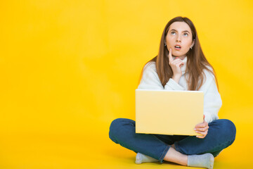 Portrait of satisfied female with beautiful smile enjoying watching movie in silver computer and sitting in lotus pose on the floor over yellow wall