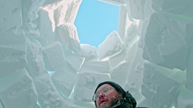 satisfied bearded man in glasses stands inside igloo of white snow blocks under clear blue winter sky low angle shot