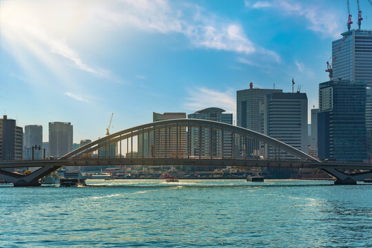 Water buses sailing on the Sumida river under the arch bridge of Tsukiji Ohashi crossed by Kanni Street with the skyscrapers of the Takeshiba district in the background.