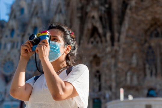young woman wearing protective mask taking pictures with camera and doing tourists things in front of landmark monuments in barcelona, spain. Tourism faces challenges due to the coronavirus pandemic