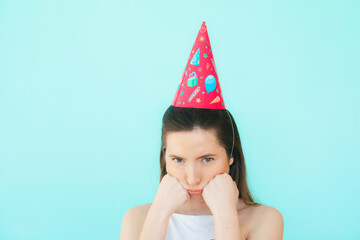 Obraz na płótnie Canvas Beautiful dissatisfied upset young sad woman in white clothes, birthday party hat, celebrating bad holiday alone on blue background isolated for advertisement