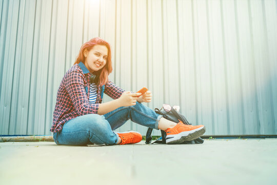Beautiful modern smiling young female teenager in a checkered shirt and jeans with headphones and smartphone sitting on the street sidewalk. Modern teens in digital world concept image