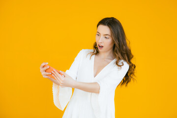 Portrait of playful attractive woman in white dress holding blank screen mobile phone and winking isolated over yellow background