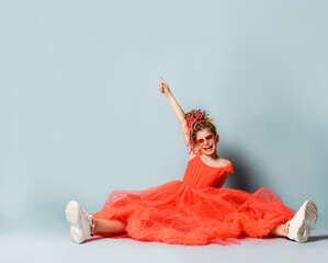 Fototapeta premium Smiling girl with colorful dreadlocks in coral dress, sneakers and suglasses sitting on floor and feeling excited with hand raised