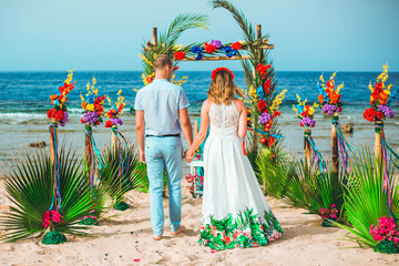 Obraz na płótnie Canvas Newlyweds go to the wedding arch. Wedding ceremony on the beach. Tropical style wedding.