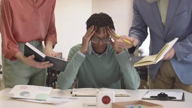 Lockdown of stressed young African man is sitting at desk in the office and listening to two unrecognizable managers accusing him