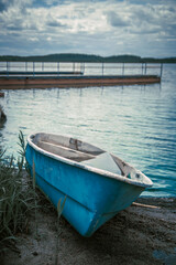 A turquoise boat lies on its side on the shore of a pond next to tall grass. Blurred lake background