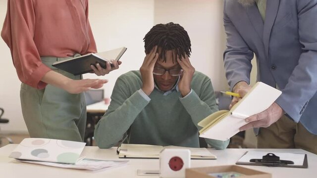 Lockdown of tired young African man sitting at desktop in the office and listening to two unrecognizable managers accusing him. Then he is looking at camera