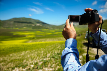 Man with a camera taking a video of panorama towards Castelluccio di Norcia (June 2020), with fields in lavish color for the beginning of flowering.