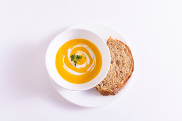 Tigela branca com creme de cenoura no prato branco mais peda&ccedil;o de p&atilde;o integral sobre a mesa branca