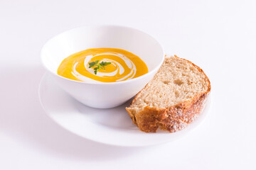 Tigela branca com creme de cenoura no prato branco mais pedaço de pão integral sobre a mesa branca