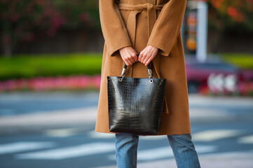 Fashionable young woman in black cowboy boots, blue jeans, beige coat, white t-shirt and crocodile print handbag in hand on the city street. Street style.