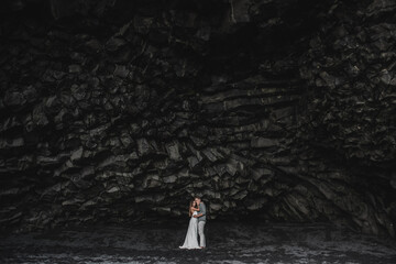 Fototapeta na wymiar Wedding in Iceland. A young guy and a girl in a white dress and a red sweater are hugging near the black rock