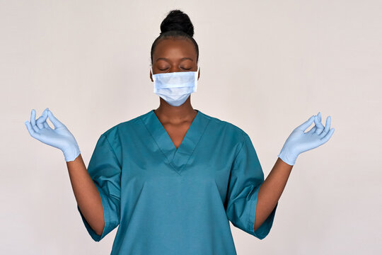 Tired Stressed Female African American Scrub Frontline Nurse Wear Face Mask Blue Uniform Meditating On Grey Background. Black Woman Doctor Eyes Closed Breathing Doing Yoga Feels Stress Relief Concept.