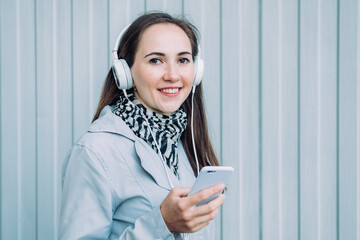 Beautiful caucasian girl in a gray coat listens to music on headphones through a smartphone