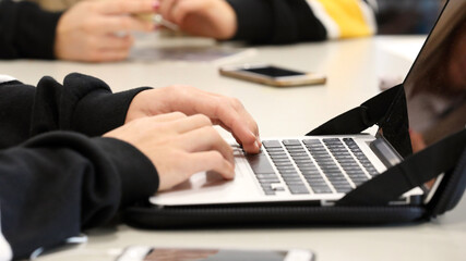 The modern learner. A student in a high school classroom environment typing on a computer or laptop keyboard with phones nearby. Deliberate shallow depth of field, fingers in focus