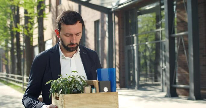 Man in 30s leaving work while getting faired and carrying box with his stuff. Unhappy male in suit remembering work moments and looking at office windows while walking at street