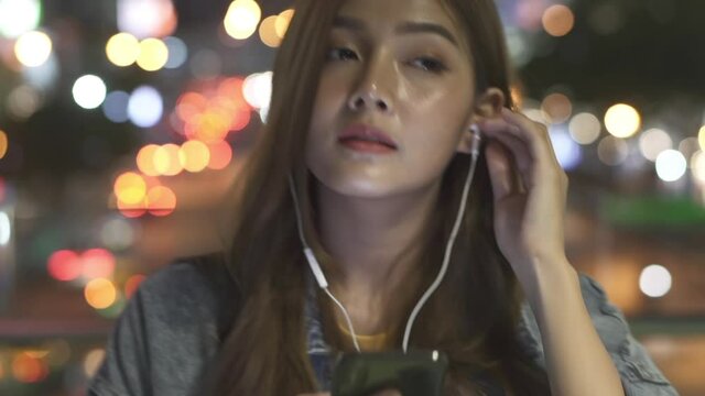 Slow motion of smiling young beautiful Asian woman girl walking in the city at night with headphones and listening online music from smartphone and enjoy looking at illuminated city night lights.