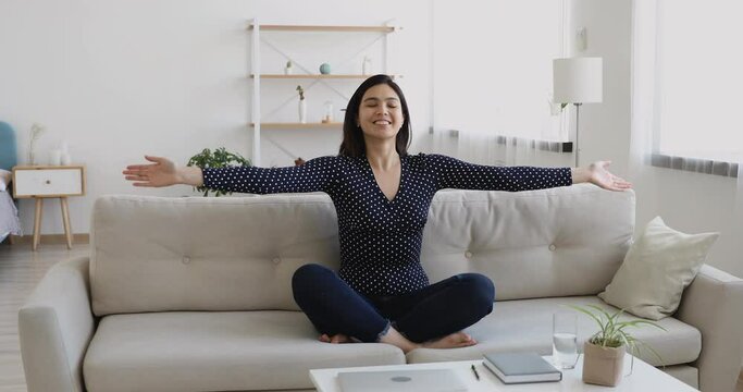 Happy motivated vietnamese young woman closed eyes seated cross-legged on comfy couch in cozy living room alone, raises and lowers arms breathing deeply do yoga exercise work out at home feels healthy