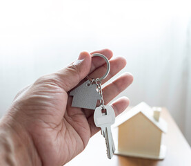 Close-up of hand holding silver keys house with house shaped keychain and home mockup on wooden table background.