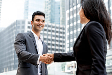 Smiling handsome young Indian businessman making handshake with businesswoman in the city with office buildings in background
