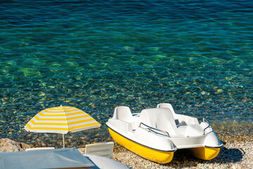 White catamaran on the pebbles beach with yellow sun umbrella, azure crystal clear sea coast. Copy space. The concept of an travel, relax, active and healthy life in harmony with nature.