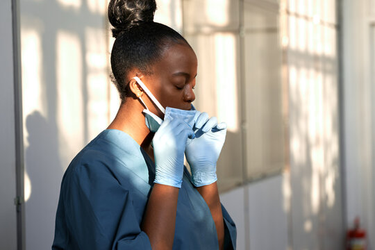 Tired female african scrub nurse wear blue uniform gloves taking off face mask to breathe in hospital. Exhausted black doctor feels stress relief concept, burnout fatigue at difficult work in hallway.