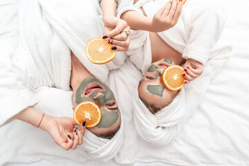 mother and daughter in white bathrobes applying pieces of orange to their eyes, in a white bath towel