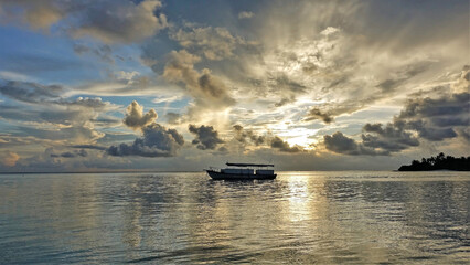 Obraz premium Amazing Maldives sunset. On a blue sky, golden clouds in the rays of the setting sun. On a glittering mirror surface of the ocean is a boat. The outlines of palm trees on an island in the distance.