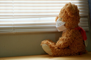 Teddy bear wearing a face mask, sitting and looking out the window with loneliness and Sadness. Coronavirus Covid-19 and pm2.5, stay at home, quarantine concept.